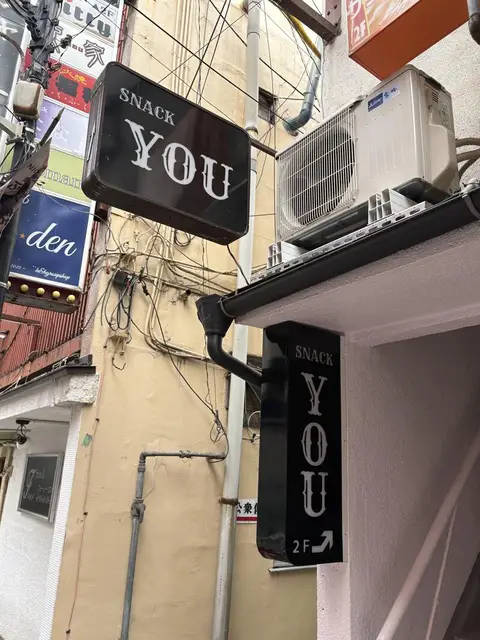 黒い箱看板に白文字を置いた「SNACK YOU」の2階案内